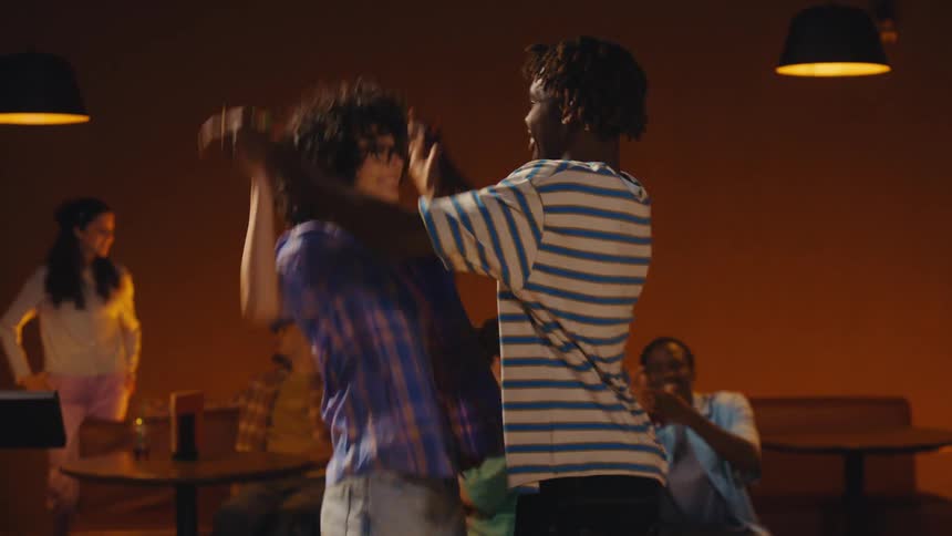 A young Black man celebrates a successful bowling shot, dancing joyfully with his friend as their group cheers in the background