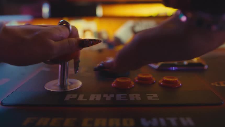 A close-up of two players with stylish, long nails energetically playing a classic retro arcade game