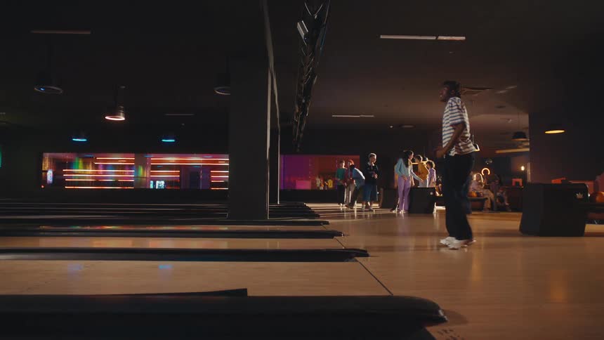 A young man throws a bowling ball in slow motion while his friends cheer him on at the bowling alley