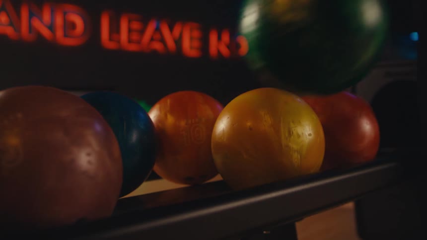 A close-up slow-motion shot of a hand selecting a green bowling ball from a rack at a bowling alley