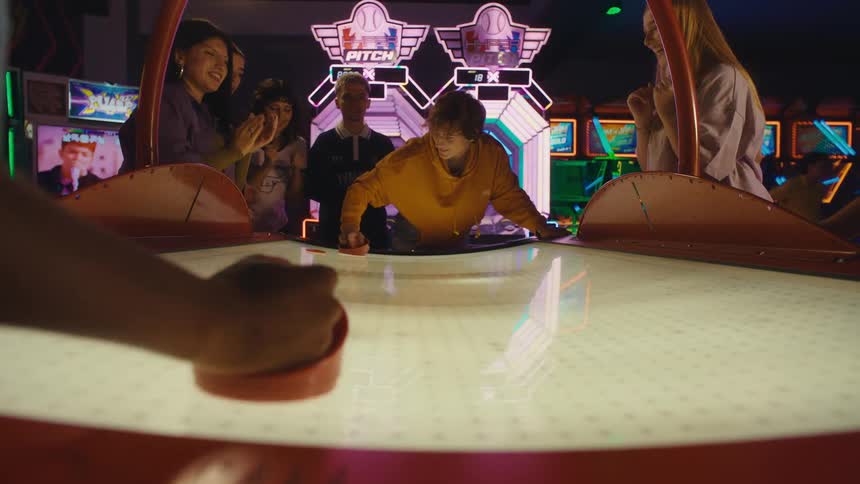 A player's point-of-view of scoring the winning goal in an intense game of air hockey as friends cheer in slow motion at a neon arcade