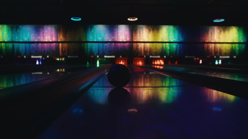 Slow motion shot captures a bowling ball rolling down a lane illuminated by colorful neon lights before falling into the gutter