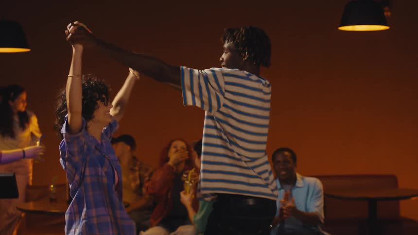 A focused young man celebrates a successful bowling throw by joyfully dancing with a friend in a retro-style alley