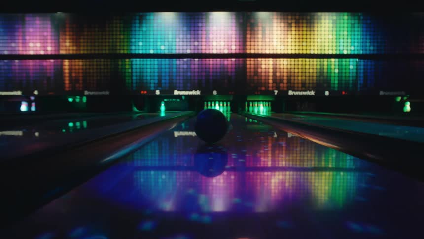A bowling ball rolls down a vibrant, neon-lit lane toward the pins in a dark, colorful alley