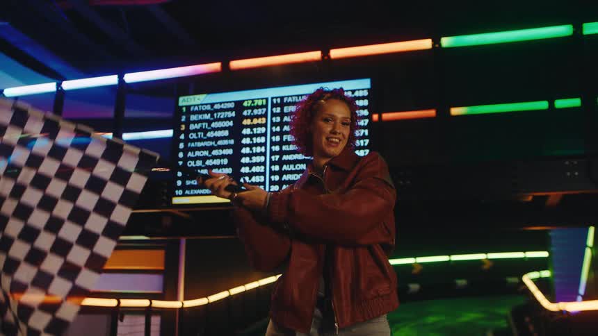 A smiling woman excitedly waves a checkered flag in slow motion at a neon-lit indoor go-kart track