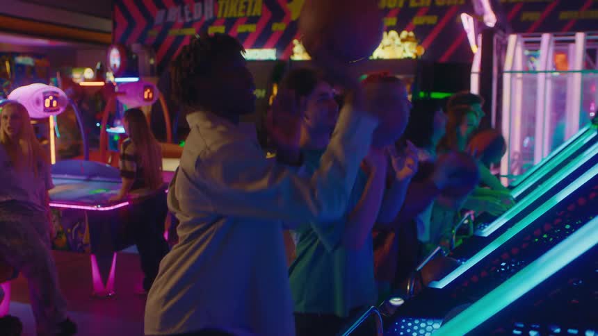 A smiling man high-fives his friend after making a basket while playing a basketball arcade game in a lively, neon-lit arcade