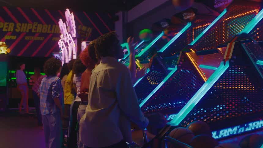 A diverse group of friends laugh and compete while playing a vibrant, neon-lit arcade basketball game