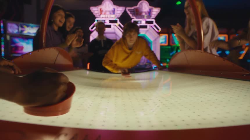 A group of friends cheer on two young men playing a competitive game of air hockey in a brightly lit arcade