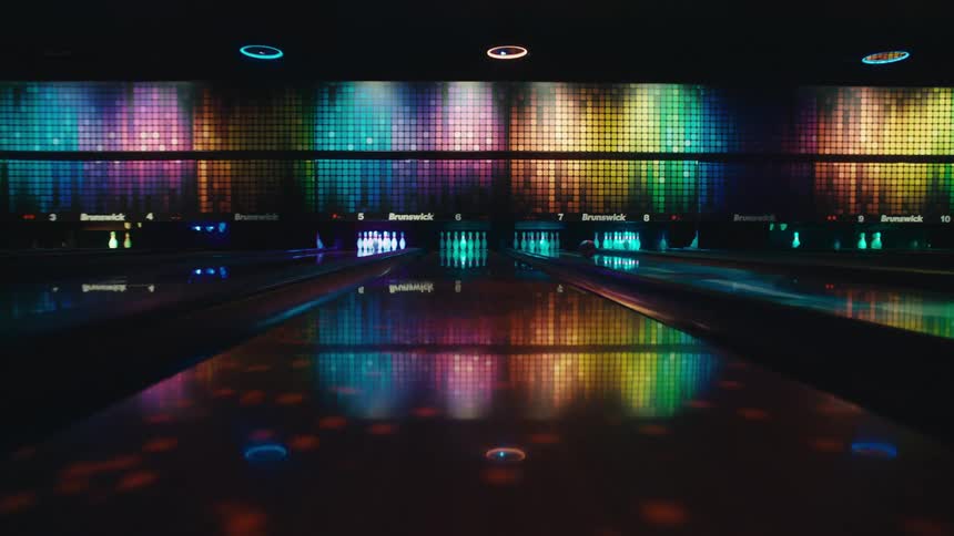 A bowling ball rolls down a futuristic, neon-lit lane and strikes the pins in a modern bowling alley