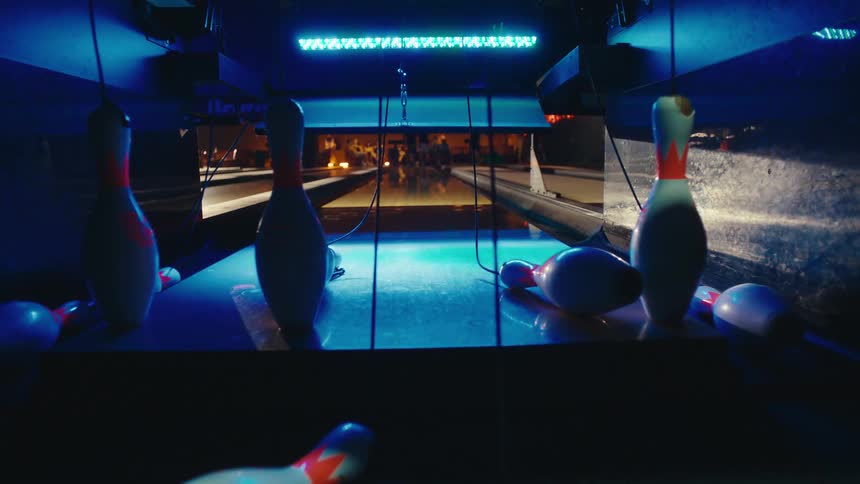 Slow motion view from behind bowling pins as a bowling ball hits for a strike and the pinsetter resets the lane under colorful neon lights