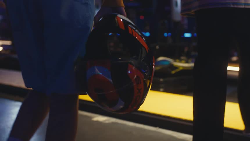 Two friends carry racing helmets as they walk towards a neon-lit indoor go-karting track in slow motion