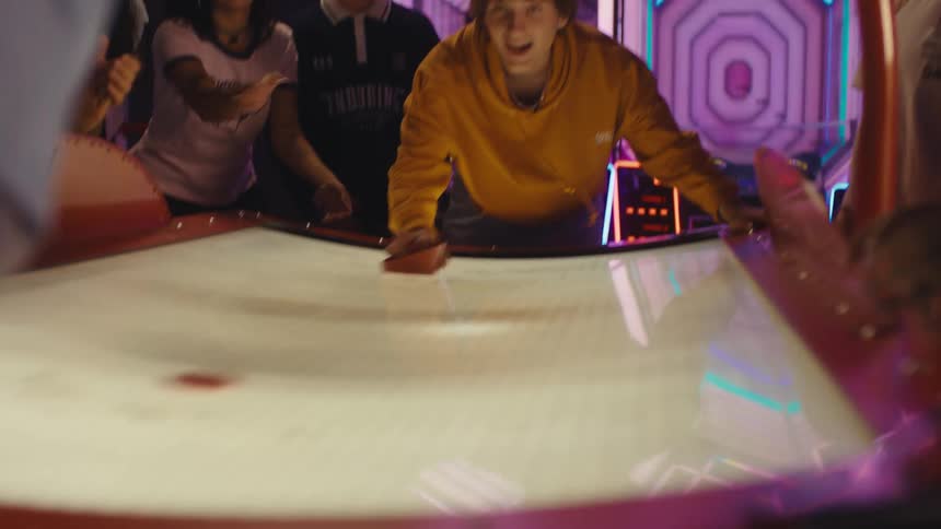 A group of happy friends cheer on a young man playing a competitive game of air hockey at a neon-lit arcade