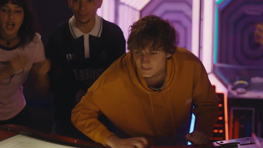 A young man intensely plays air hockey while his friends enthusiastically cheer him on in a neon-lit arcade