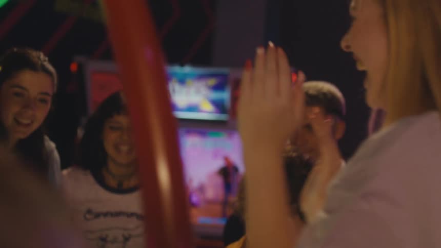 A group of excited friends cheer and dance while watching an intense game of air hockey at a vibrant arcade