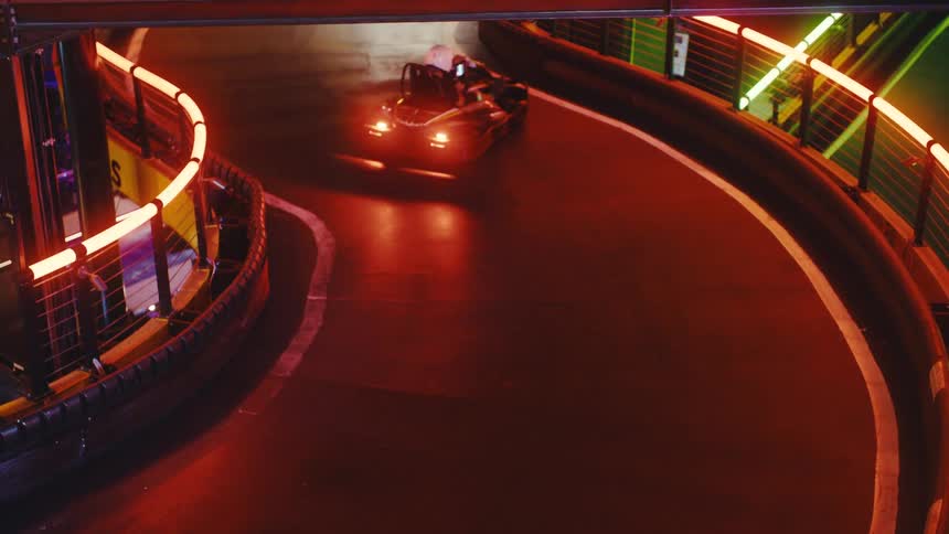 Go-karts race around a tight, neon-lit corner on an indoor track before one car spins out