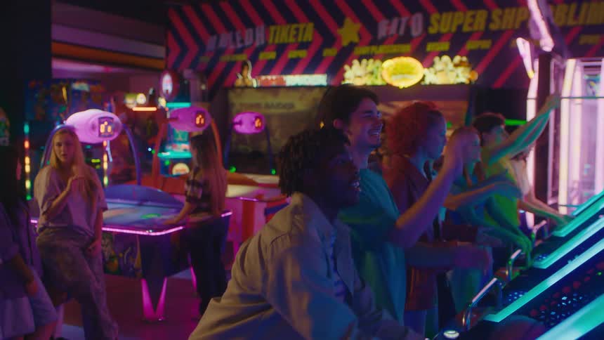A man high-fives his friend while a diverse group of young people enjoy a competitive basketball game at a neon-lit arcade