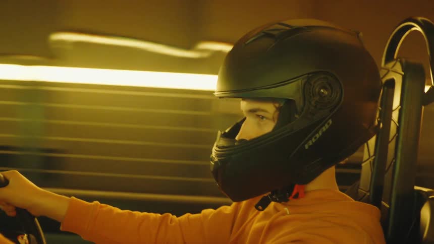 Profile shot of a focused young man in a helmet driving a go-kart on an illuminated indoor track
