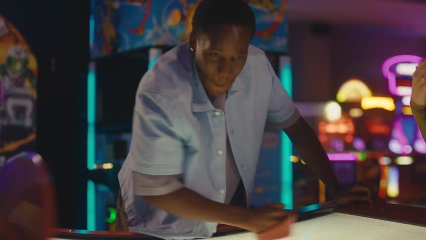 A smiling young man energetically plays a game of air hockey with a friend in a vibrant, neon-lit arcade