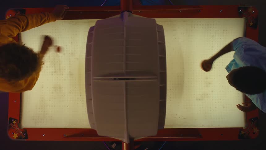 A top-down view captures a fast-paced and competitive game of air hockey between two friends in a neon-lit arcade