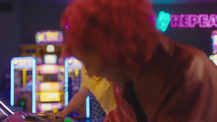 A stylish young woman with red hair smiles and laughs while competing against a friend on a classic arcade game in a vibrant, neon-lit gaming hall