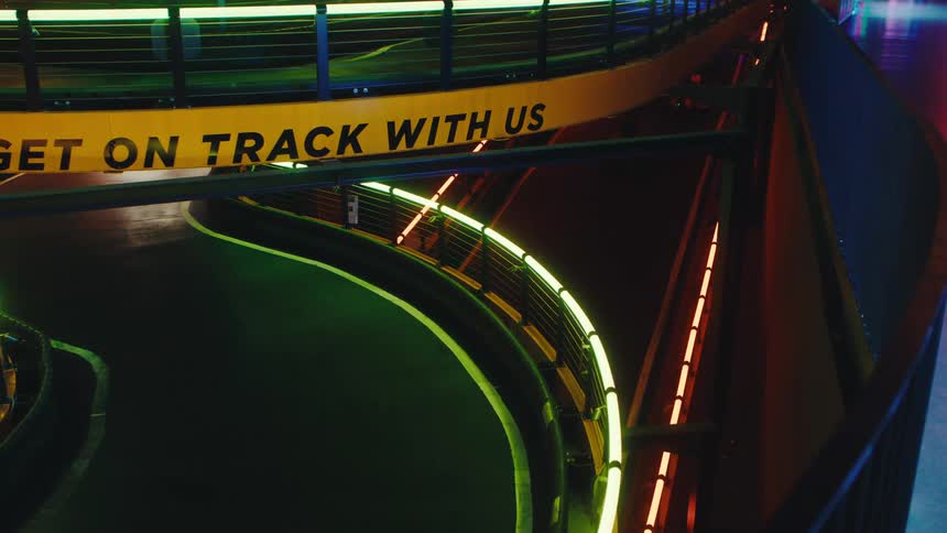Racers in go-karts speed along a futuristic indoor track with colorful neon lighting