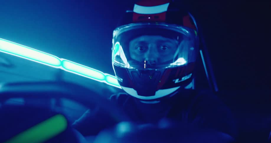 A focused racer's face is illuminated by colorful neon lights reflecting in their helmet's visor as they drive a go-kart