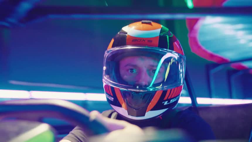 An intense go-kart driver wearing a helmet races through a track illuminated by vibrant blue and purple neon lights