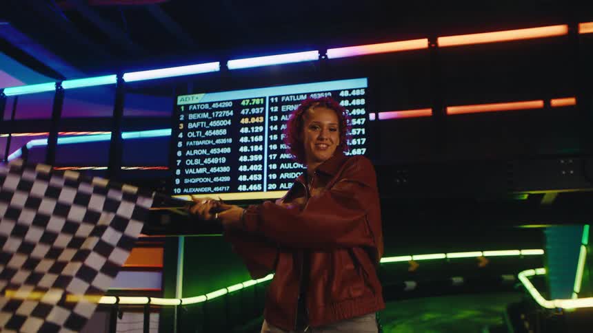 A happy young woman with red hair excitedly waves a checkered flag at a go-kart race track in slow motion
