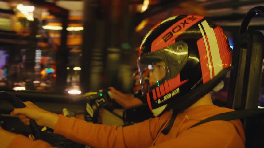 Close-up side view of a person wearing a helmet while driving a go-kart on a vibrant, neon-lit indoor race track
