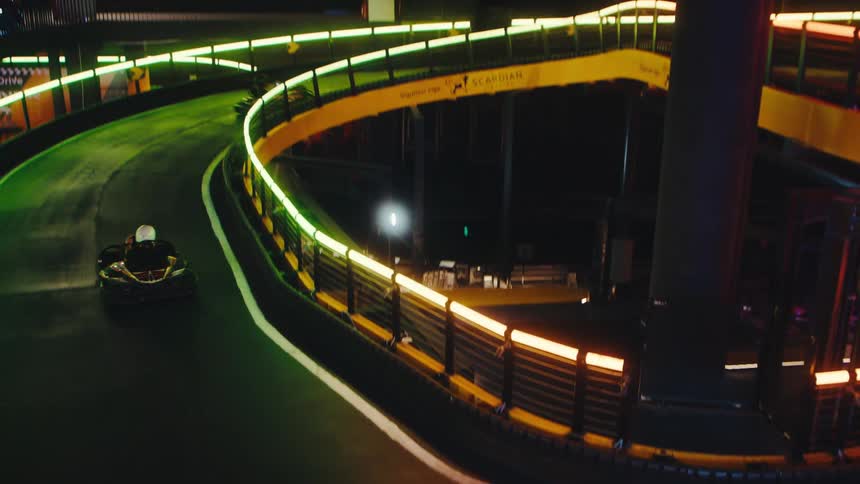 Go-kart racers speed around a multi-level indoor track illuminated by vibrant neon lights