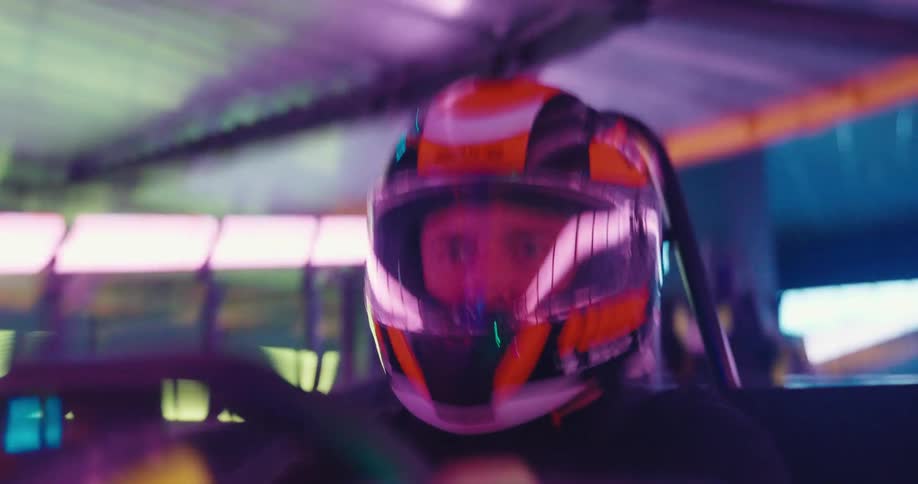 Close up of a focused man in a helmet driving a go-kart in slow motion on a colorful neon-lit indoor track