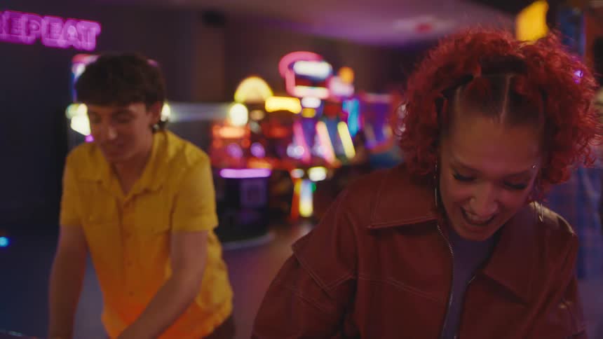 An energetic woman with curly red hair laughs and cheers while playing a competitive game against a friend in a vibrant arcade