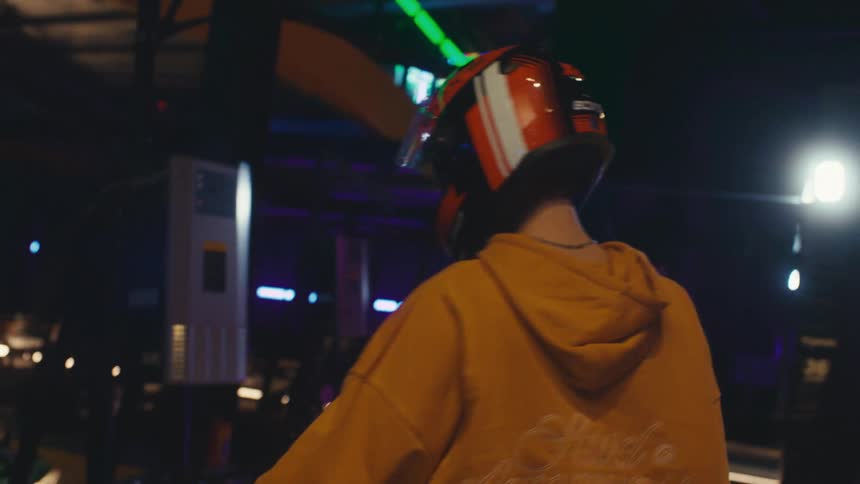 A young person in a yellow hoodie puts on a racing helmet and walks towards their go-kart at a vibrant, neon-lit indoor track