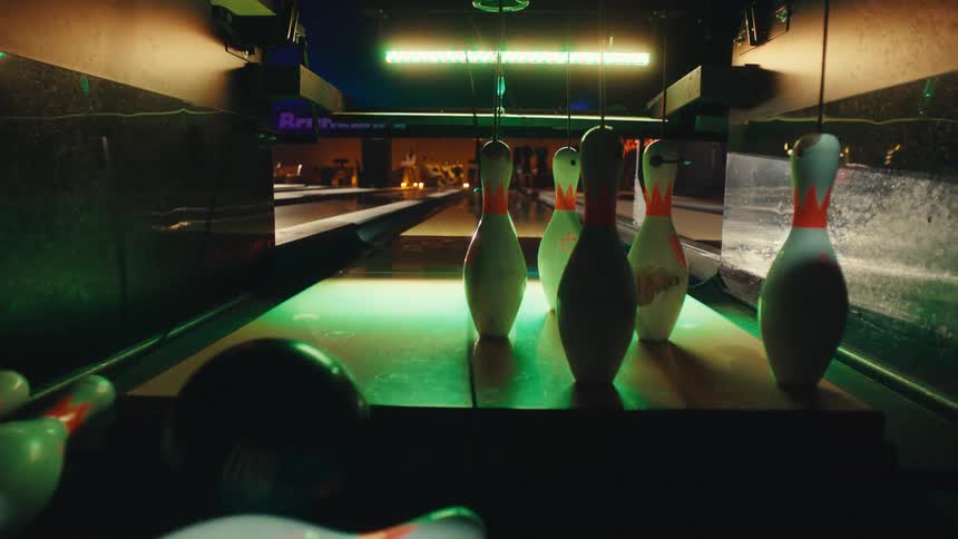 A dramatic slow-motion shot from the pinsetter's perspective captures a bowling ball hitting a spare, sending pins flying as the alley lights change color