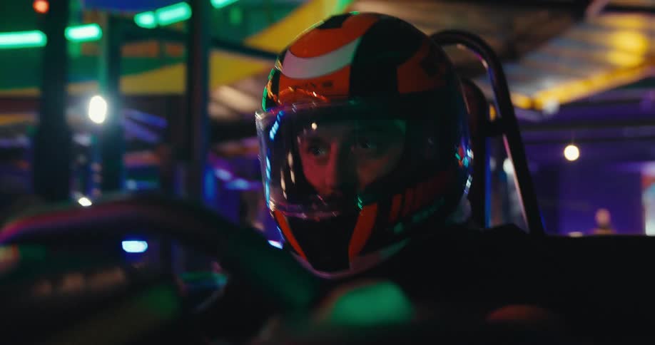 Intense close-up of a determined man in a helmet racing a go-kart on a vibrant, neon-lit indoor track