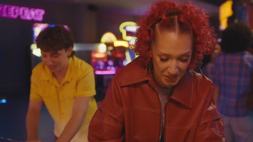 A young woman with curly red hair smiles and laughs while playing an intense game of air hockey with a friend in a neon-lit arcade