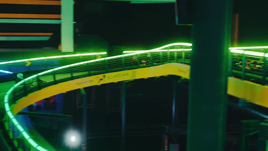 Friends race go-karts at high speed on a winding, multi-level indoor track illuminated by vibrant green neon lights