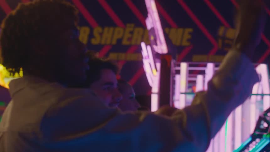 A diverse group of friends laugh and compete while playing a basketball arcade game under neon lights