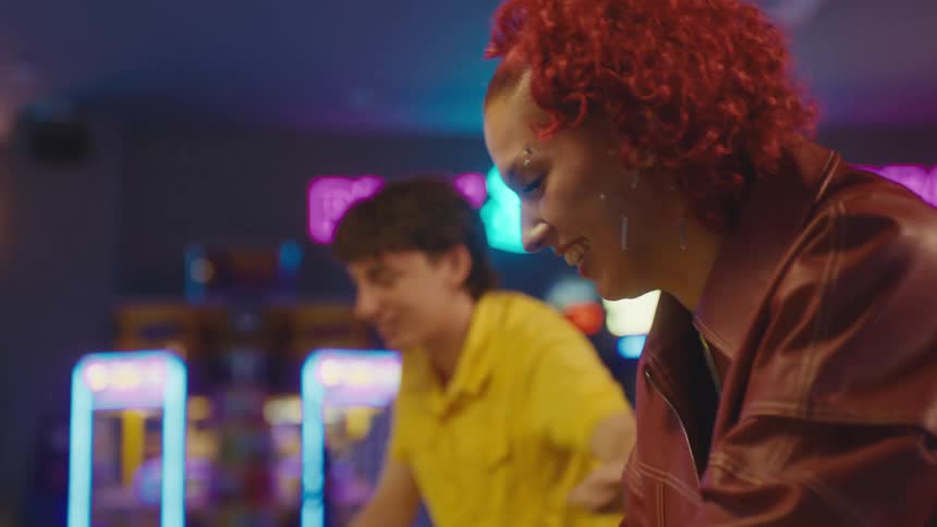 A young woman with red hair and her friend laugh and play a retro arcade video game in a neon-lit amusement park