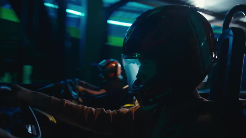 A close-up side profile shot of a go-kart driver wearing a helmet while racing around a neon-lit indoor track at night