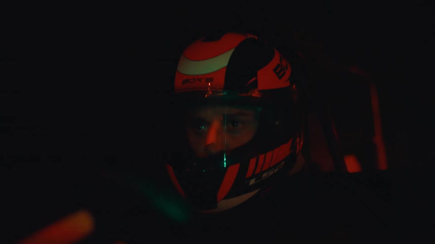 A focused driver in a racing helmet navigates a go-kart track at night, with dynamic, colorful lights flashing across his visor