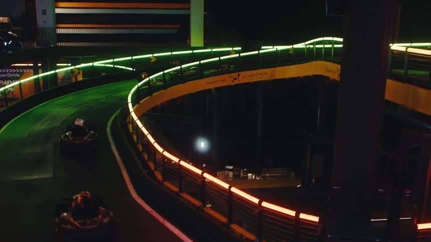 High-angle view of people racing go-karts on a winding indoor track illuminated by colorful neon lights at night