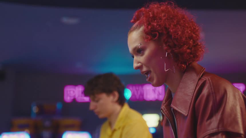 A young woman and man smile and laugh while playing a classic competitive video game at a retro arcade