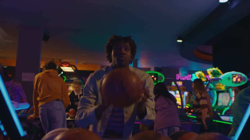 A young man concentrates while playing a basketball shooting game in a vibrant, neon-lit arcade