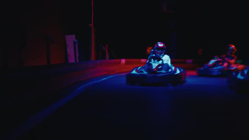 A thrilling first-person view of a go-kart race on a multi-level indoor track with vibrant, changing neon lights