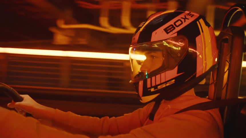 A racer wearing a helmet drives a go-kart at high speed on a colorful, neon-lit indoor track