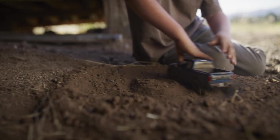 A young boy in a cowboy hat finds a shady spot under a wooden structure to play with his toy truck in the dirt