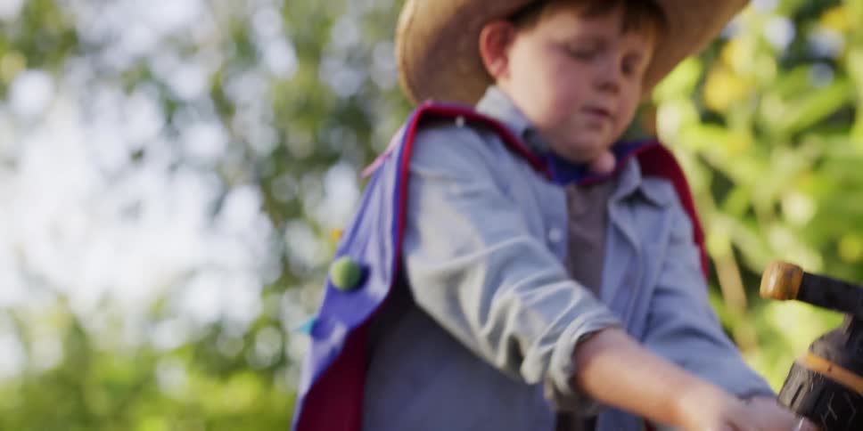 A young boy wearing a cowboy hat and superhero cape pours water over his blue toy truck while playing outside in slow motion
