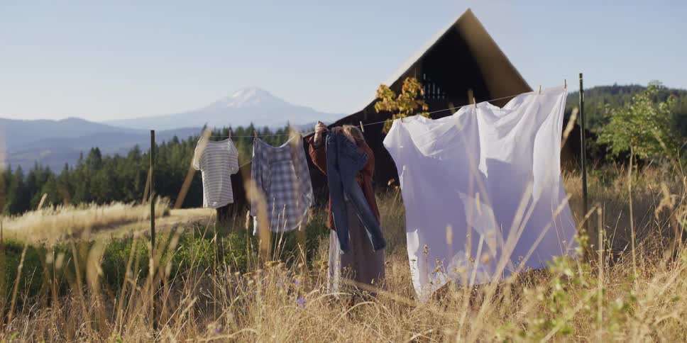 A woman in slow motion hangs jeans on a rustic clothesline in a sun-drenched field with a mountain view