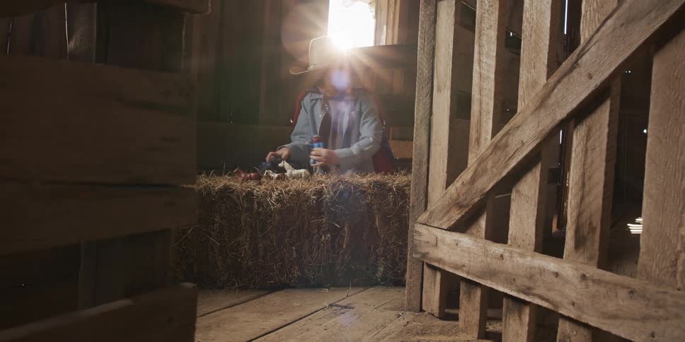 Sunlight streams into a rustic barn as a young boy in a cape and cowboy hat plays with toys on a bale of hay